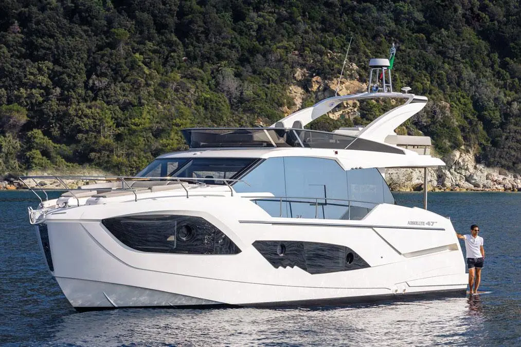 A sleek white luxury yacht, the Absolute 47, floating in calm waters with a man standing on the deck against a green hillside.